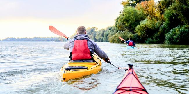 Three kayakers paddle their brightly-coloured recreational kayaks near the water's edge.